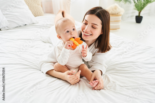 Baby chews on a teething toy while sitting on the bed with mom.