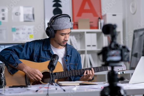Young Asian male graphic designer blogger influencer playing guitar while shooting education tutorial vlog training filming video course for social media at studio.