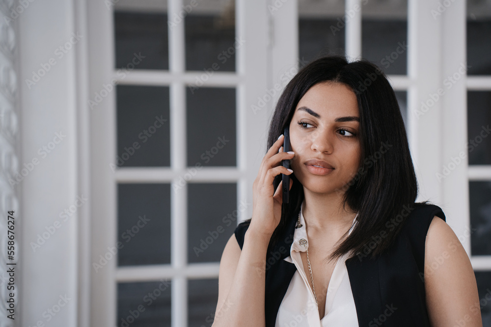 Adorable calm African American young woman talking by phone looks aside in disbelief standing at home. Beautiful Brazilian girl using cellphone at office. Confident businesswoman talks with partner. Adorable calm African American young woman talking by phone looks aside in disbelief standing at home. Beautiful Brazilian girl using cellphone at office. Confident businesswoman talks with partner.