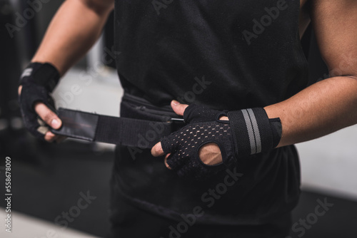 Bild auf Leinwand An anonymous man wearing and tank top and training gloves tightening the velcro weightlifting belt on his waist at the gym