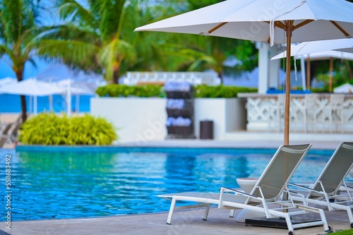 White sand beach and blue sea landscape view of a tropical resort hotel beside modern swimming pool, white parasols, palm trees, deck beach chairs in the golden sunset time