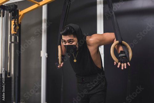 Fototapeta Naklejka Na Ścianę i Meble -  A masked asian man leaning on wooden still rings. A gymnast preparing to train at the gym.