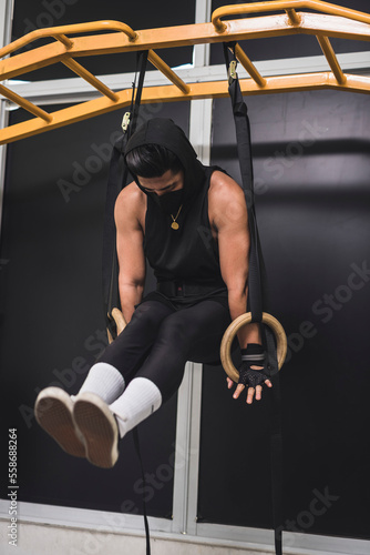 A hooded man doing an L-sit exercise on wooden still rings at the gym. Calisthenics exercises on gymnastics rings.