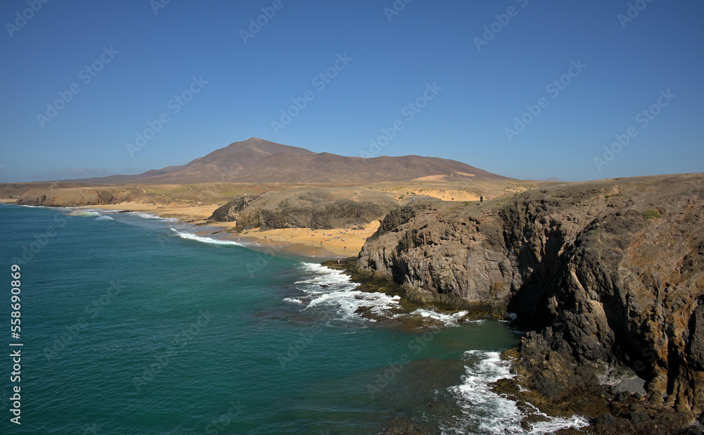Eine Küstenlandschaft mit Strände aus Sand und badende Menschen und im Hintergrund ist bei blauem Himmel ein Berg.
