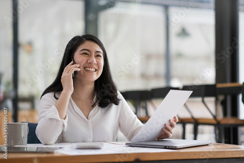 Happy smiling asian business woman working on laptop at office, using smart phone. Businesswoman sitting at her working place