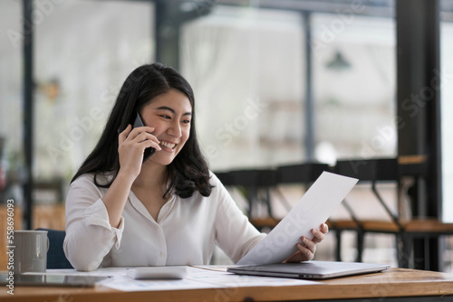 Happy smiling asian business woman working on laptop at office, using smart phone. Businesswoman sitting at her working place