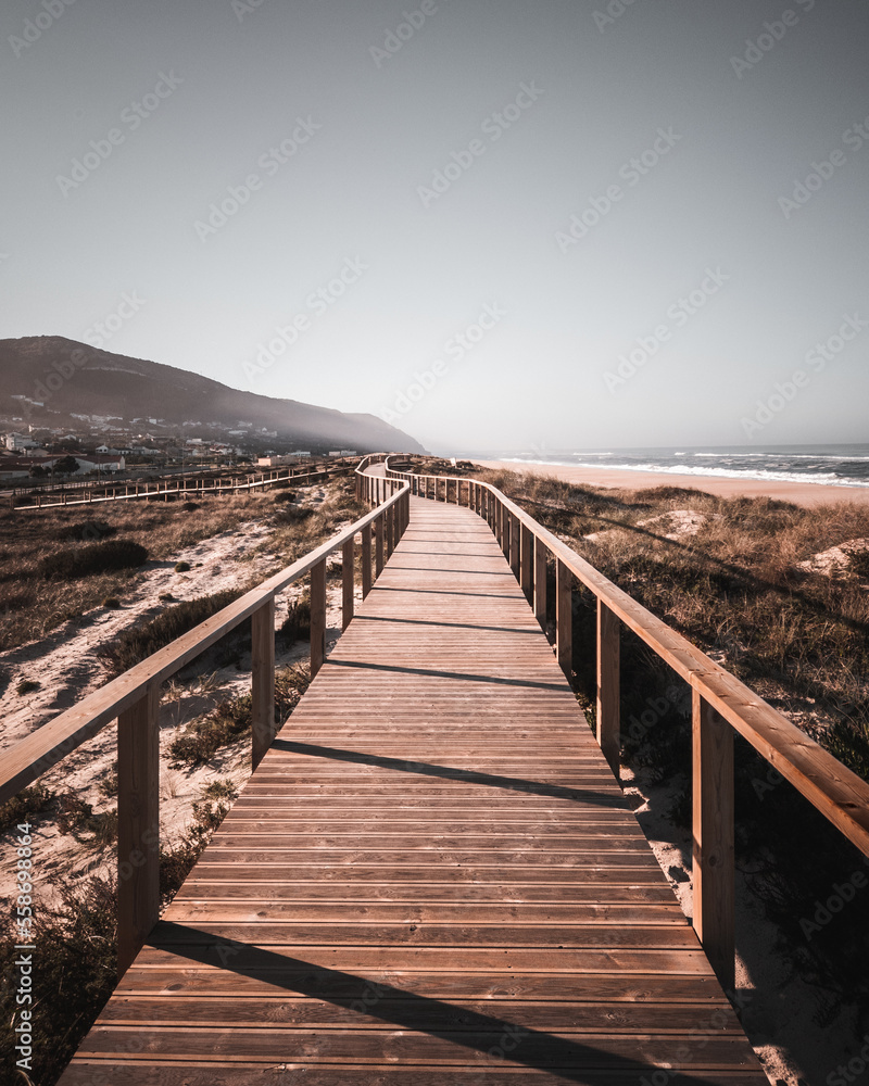 A long boardwalk on a grassy sand dune at the beach leading to the mountain and ocean with desaturated colors. The way ahead concept, Quiaios Beach, Portugal