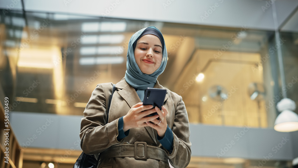 © Gorodenkoff - Confident Female Team Manager wearing Hijab Answering Emails on Smartphone After Receiving Good Performance Report. Portrait of a Young Muslim Woman Walking in a Hallway of an Office and Smiling.