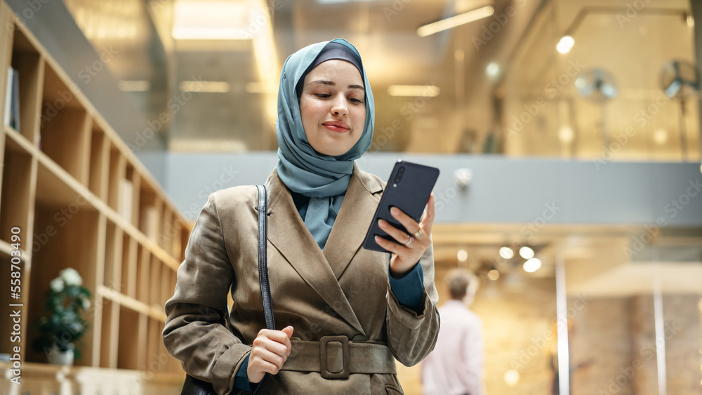 Muslim Woman wearing Hijab Checking Email App on her Smartphone After a Succesful Meeting. Portrait of a Female Content Manager Walking Through a Busy Workplace Hall. Low Angle Muslim Woman wearing Hijab Checking Email App on her Smartphone After a Succesful Meeting. Portrait of a Female Content Manager Walking Through a Busy Workplace Hall. Low Angle