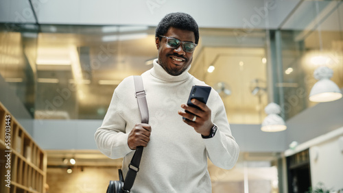 Portrait of Young Black Man Feeling Confident as He Walks Smiling and Checking His Phone, in a Busy Office Hallway. He is Browsing the Internet on His Smartphone. Low Angle