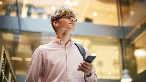 Portrait of Young Caucasian Man Feeling Confident as He Walks Smiling and Checking His Phone, in a Busy Office Hallway. He is Browsing the Internet on His Smartphone. Low Angle