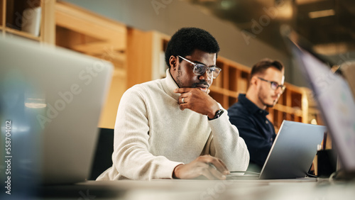 Portrait of Black Creative Young Man Arriving at Office for Work Office and Working on Laptop. Supervisor Smiling While Answering Email Questions From Colleagues. People Working in the Background