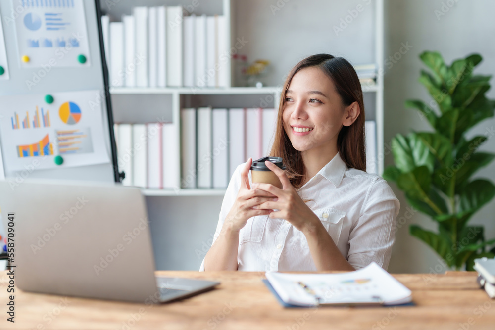 Pretty Asian businesswoman sitting happily working on her laptop and drinking coffee smiling and laughing brightly in the office. Pretty Asian businesswoman sitting happily working on her laptop and drinking coffee smiling and laughing brightly in the office.