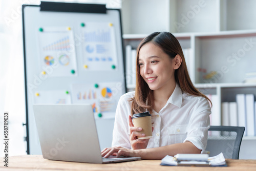 Pretty Asian businesswoman sitting happily working on her laptop and drinking coffee smiling and laughing brightly in the office.