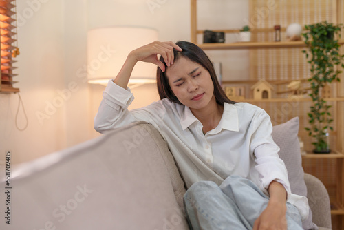 Young Asian woman suffering from stress from an existing disease having a headache. Migraine sitting alone on the sofa at home frustrated. Desperate Asian woman feeling lonely, tired.
