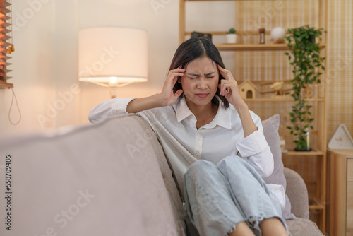 Young Asian woman suffering from stress from an existing disease having a headache. Migraine sitting alone on the sofa at home frustrated. Desperate Asian woman feeling lonely, tired.