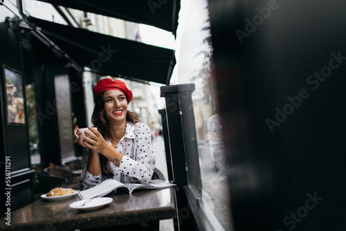 Young stylish woman in red beret having a french breakfast with coffee and croissant sitting oudoors at the cafe terrace. Fresh Baked Croissants. 