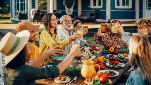 Family and Multiethnic Diverse Friends Gathering Together at a Garden Table Dinner. Old and Young People Raising and Clinking Glasses with Fresh Orange Juice and Celebrating the Occasion.