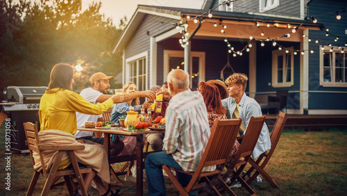 Family and Multiethnic Diverse Friends Gathering Together at a Garden Table Dinner. Old and Young People Toasting and Clinking Glasses with Fresh Orange Juice and Celebrating an Event.