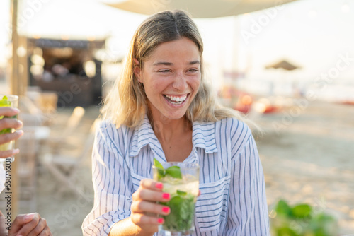 A young blond girl celebrating in chiringuito clinking glasses with mojito - People and lifestyle concept photo