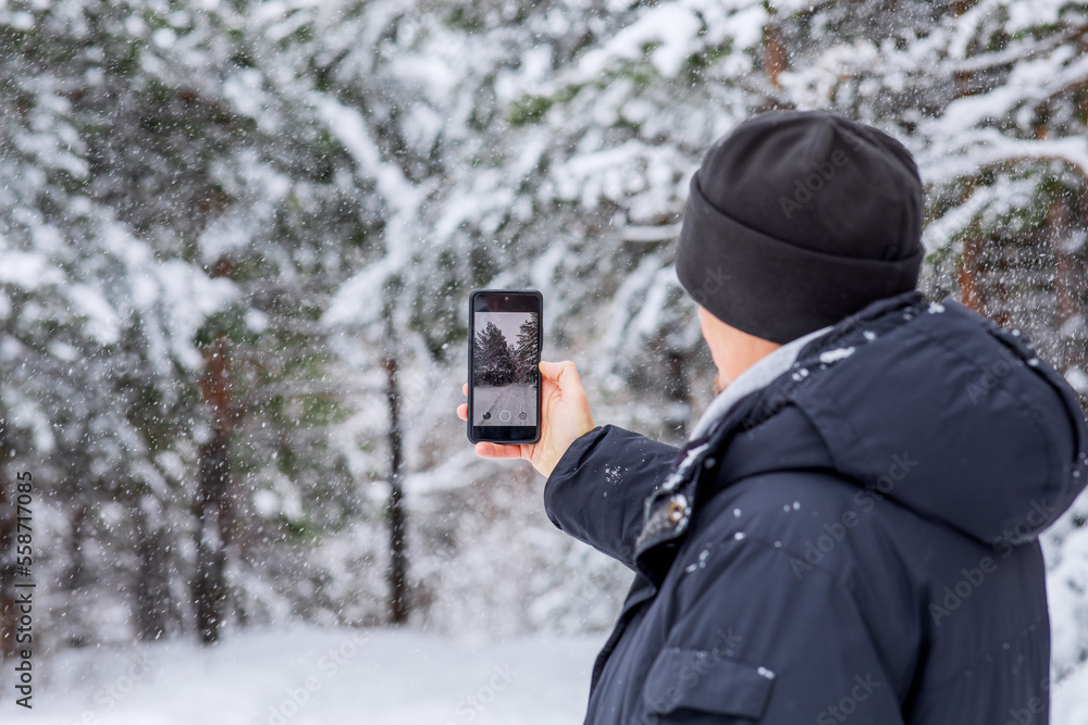 A man takes a picture of a snow-covered pine forest on his phone. Winter sports walk. A man takes a picture of a snow-covered pine forest on his phone. Winter sports walk.
