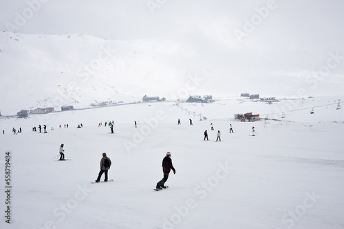 Skiers go downhill skiing on snow-covered mountain ski slope against the backdrop of a cloudy sky and ski lifts. Winter. Extreme sport and travel content  