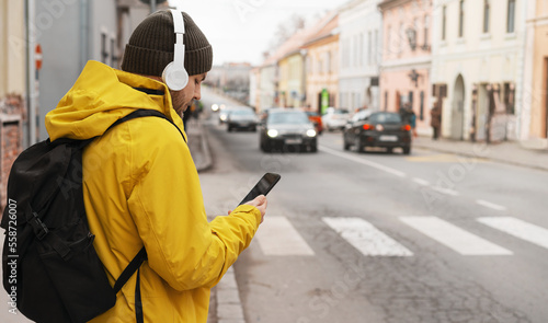 Man in yellow jacket and headphones standing next to road in city using his smart phone while waiting taxi.