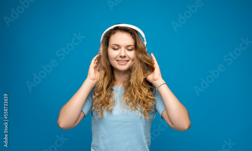 Portrait of charming curly hair woman with a closed eyes in blue t-shirt in massive white headphones listening to her favourite music band on blue background.