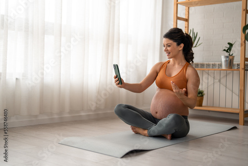 Cheerful pregnant woman sitting on the floor in lotus position and having a video call.