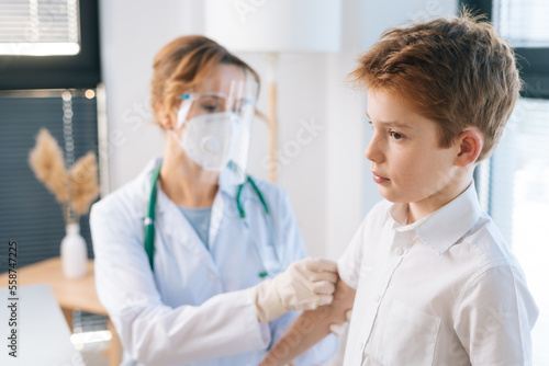 Female doctor wearing white uniform and safety mask vaccinating cute child boy at hospital with modern light interior. Concept of vaccination program  prevention of infectious diseases.
