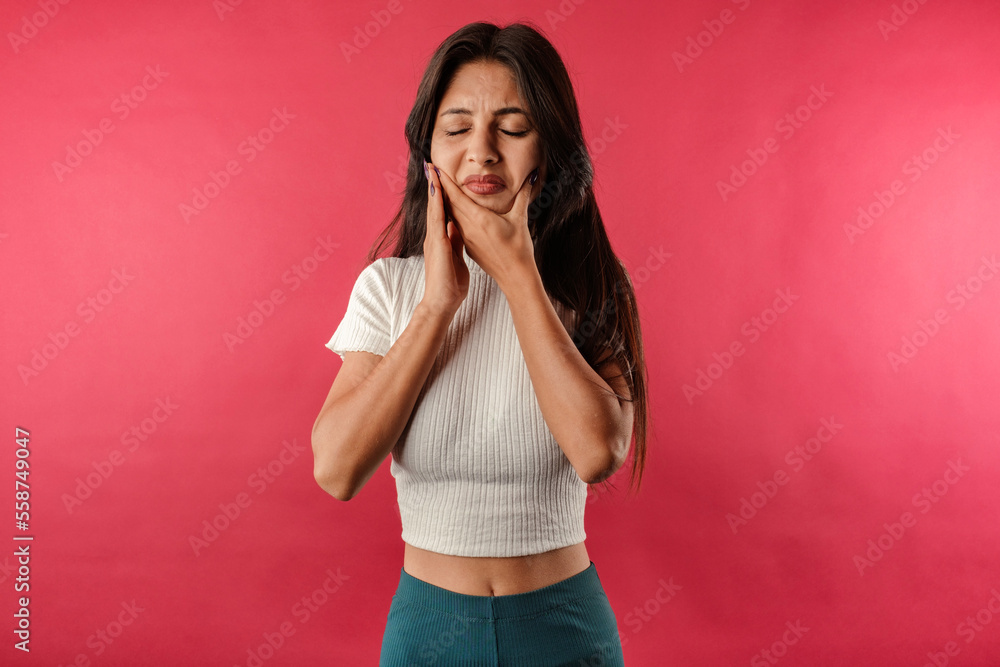 Brown-haired woman wearing white ribbed crop isolated over red background touching cheek closed eyes, suffering from a toothache. Tries to get relief by touching chin. Dentist concept. Brown-haired woman wearing white ribbed crop isolated over red background touching cheek closed eyes, suffering from a toothache. Tries to get relief by touching chin. Dentist concept.