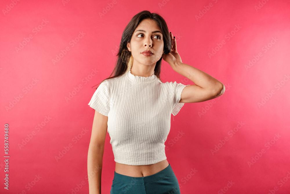 Beautiful brunette woman wearing white ribbed crop isolated over red background bewildered, eavesdropping or on what is being said in secret. Astonished eavesdrop secret rumor listen. Beautiful brunette woman wearing white ribbed crop isolated over red background bewildered, eavesdropping or on what is being said in secret. Astonished eavesdrop secret rumor listen.