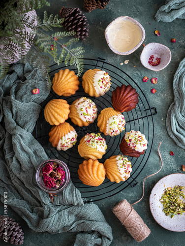 Festive madeleines on a cooling rack decorated with white chocolate, pistachios and red petals over a green background and cloth photo