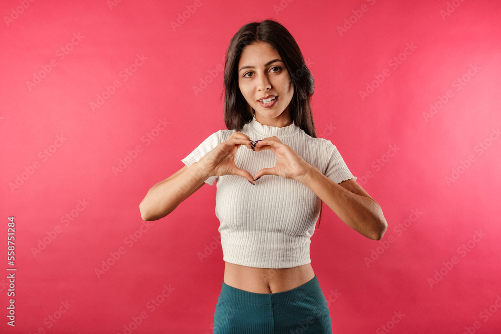 Cute brunette woman wearing white ribbed crop isolated over red background making a heart gesture with her fingers in front of her chest showing her love. With love, you are not alone. I love you. Cute brunette woman wearing white ribbed crop isolated over red background making a heart gesture with her fingers in front of her chest showing her love. With love, you are not alone. I love you.