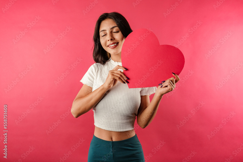 Brunette woman wearing white ribbed crop isolated over red background smiling with love in a warm, friendly pose with a large cardboard heart shape. Cuddle big paper card heart shape figure. Brunette woman wearing white ribbed crop isolated over red background smiling with love in a warm, friendly pose with a large cardboard heart shape. Cuddle big paper card heart shape figure.