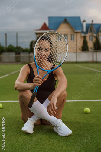 Girl with dark hair on the tennis court with a tennis racket posing in a black bodysuit