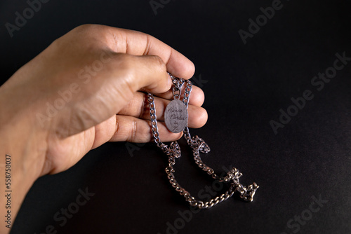 Mano sosteniendo collar con placa que tiene grabada la frase “nunca pierdas la fe” en fondo negro y blanco y negro. photo