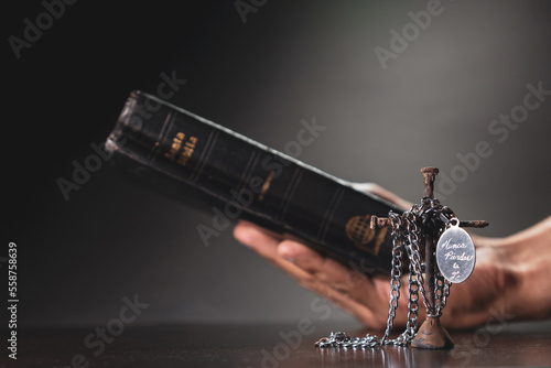 Mano en posición de orar o rezar y sosteniendo la santa biblia y cruza de clavos envuelto con un collar con placa que tiene grabada la frase “nunca pierdas la fe” en fondo negro y blanco y negro