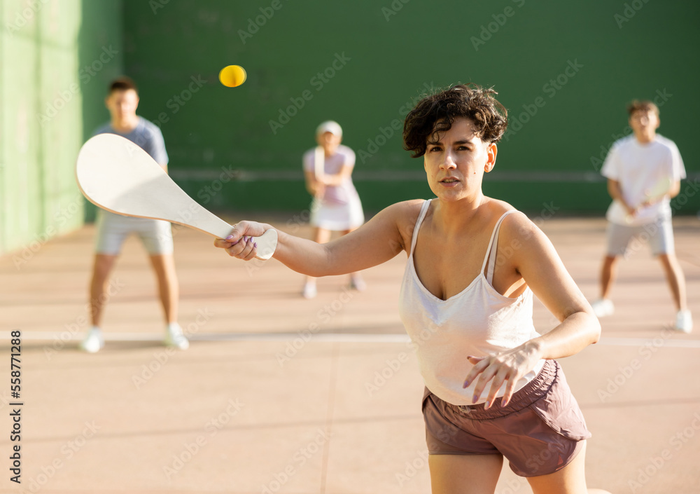 Sporty young Argentinian woman playing traditional team match of pelota at open-air fronton on summer day, ready to hit ball with wooden bat