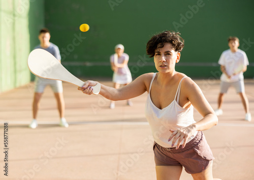 Sporty young Argentinian woman playing traditional team match of pelota at open-air fronton on summer day, ready to hit ball with wooden bat © JackF