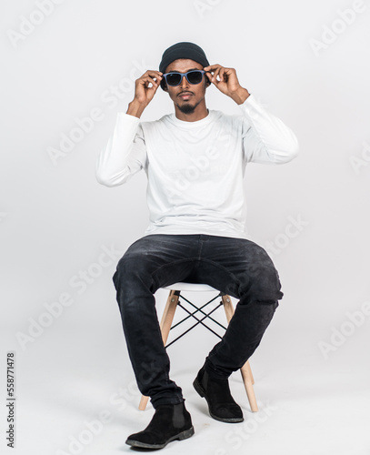 An african man wearing white blank long sleeve shirt, with a hat and glasses sitting down to a chair