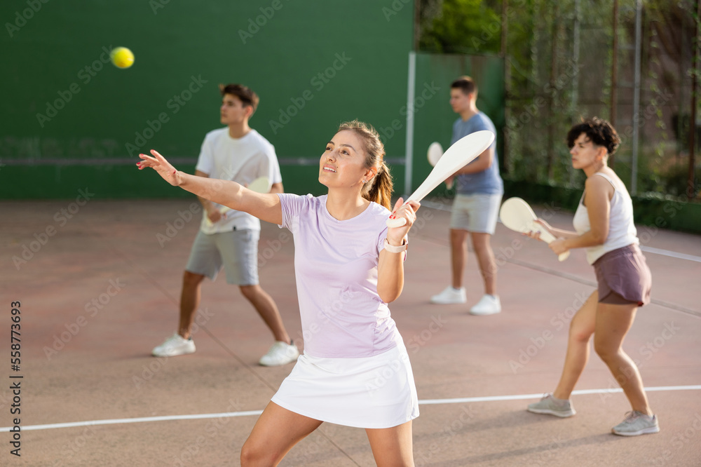 Sporty young Argentinian woman playing traditional team match of pelota at open-air fronton on summer day, ready to hit ball with wooden bat