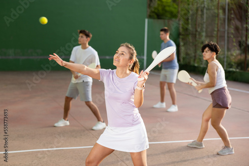 Sporty young Argentinian woman playing traditional team match of pelota at open-air fronton on summer day, ready to hit ball with wooden bat