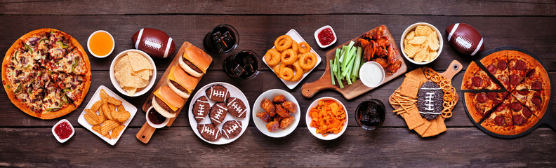 Super Bowl or football theme food table scene. Pizza, hamburgers, wings, snacks and sides. Above view on a dark wood banner background.
