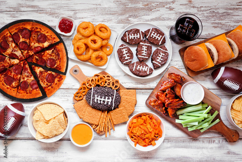 Super Bowl or football theme food table scene. Pizza, hamburgers, wings, snacks and sides. Top view on a white wood background.