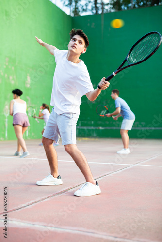 Sporty young Argentinian guy playing popular team game frontenis at open-air fronton court on summer day, ready to hit rubber ball with racquet © JackF