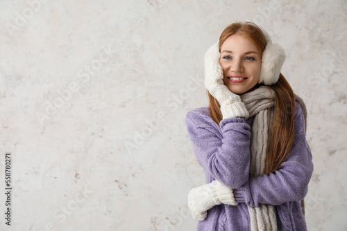 Young redhead woman in warm earmuffs and scarf on light background