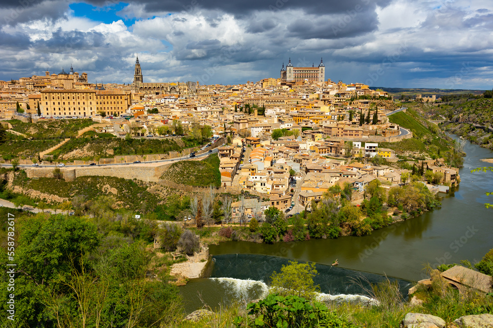 Naklejka premium View of historic area of Toledo city on banks of Tagus River overlooking belfry of Primatial Cathedral of Saint Mary and Alcazar fortress in cloudy spring day, Spain