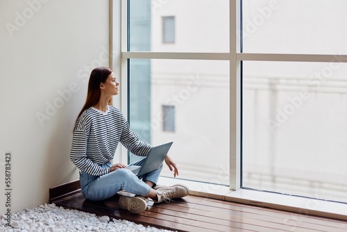 A woman freelancer sits with her laptop full-length at a large window, looking out at tall buildings in the city, work, training and shopping online