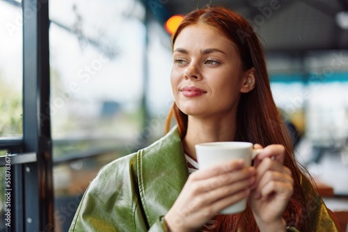 Portrait of a woman influencer drinking a drink from a mug in a cafe and smiling looking out the window, content blogger close-up